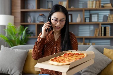 Kanepede pizza servisi yapmaktan hoşlanan Asyalı bir kadın telefonla konuşuyor, ev ortamında kendini rahat hissediyor. Tecrübe, lezzetli konforlu yiyeceklerle modern iletişimi rahat ortamlarda harmanlıyor..