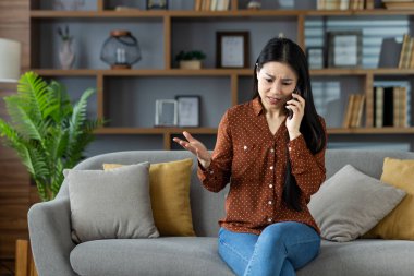 Endişeli Asyalı kadın akıllı telefon kullanıyor, koltukta oturuyor, önemli konuları tartışıyor. Modern oturma odası ortamında duygu, iletişim ve teknolojiyi gösterir.