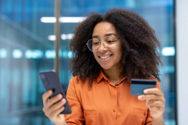İş yerindeki genç başarılı kadın ofis içi yakın plan, telefon ve banka kredi kartı olan iş kadını, online internet mağazasından satın alma, bankacılık uygulaması kullanarak para aktarma.