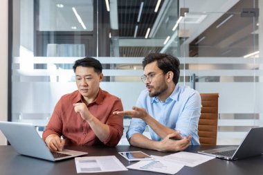 Ofis çalışanları dizüstü bilgisayara bakarak iş projesini tartışıyor, iki farklı adam bilgisayarla masada oturuyor, ciddi ve takım çalışmasına odaklanmış..