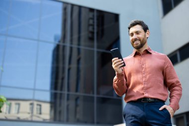 Akıllı telefonunda uygulama kullanan başarılı bir adam, iş adamı şehirde dolanıp ofis binasının dışından mutlu bir şekilde bir mesaj yazarak gülümsüyor..