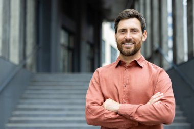 Ofis binasının dışında kendinden emin ve başarılı bir iş adamının portresi. Kırmızı gömlekli adam gülümsüyor ve kollarını kavuşturmuş kameraya bakıyor..