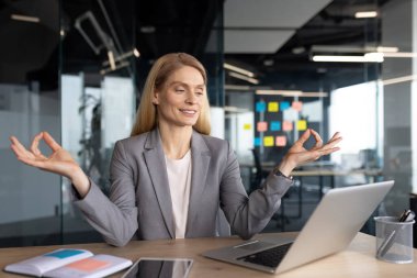 Lotus 'ta meditasyon yapan neşeli kadın iş yerinde ofiste poz veriyor. İş elbisesi giymiş olgun bir iş kadını, dinleniyor ve rahatlıyor. İnternetten yoga dersleri izliyor, dizüstü bilgisayar kullanıyor..