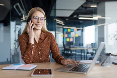 Ofisteki bir masada oturan başarılı bir kadın telefonda konuşuyor. İş yerindeki iş kadını mutlu bir şekilde gülümsüyor, dizüstü bilgisayarla çalışıyor..