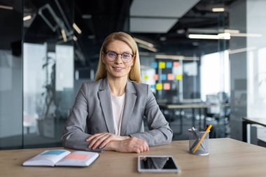 Çevrimiçi iş toplantısı, konferans. Olgun bir kadın gülümsüyor ve kameraya bakıyor. Ofis içinde belgeler ve kağıtlarla çalışıyor. Takım elbiseli bir iş kadını. Web kamera görüntüsü.