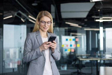 İş elbisesinde başarılı bir kadın telefonda uygulama kullanıyor. İş yerindeki iş kadını, akıllı telefondan gülümseyen, pencerenin yanında duran..