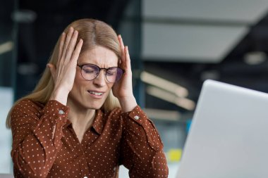 Hayal kırıklığına uğramış ve üzgün bir kadın iş yerinde el ele tutuşuyor. Ofisin içinde dizüstü bilgisayarla çalışan iş kadını, başarının sonuçlarından memnun değil.