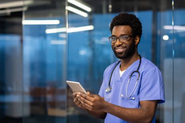 Elinde tablet bilgisayarla, gülümseyen ve kameraya bakan başarılı bir doktorun portresi. Doktor modern kliniğin içinde, akşamları çalışıyor, hastalara uzaktan danışmanlık yapıyor..