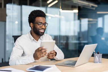 İşyerinde bilgisayarla çalışırken gülümseyen başarılı bir işadamı elinde tablet bilgisayarla elinde memnuniyet dolu bir adam, genç müteahhit, yatırımcı.