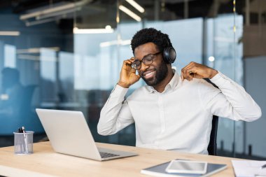 İş yerindeki genç adam dizüstü bilgisayarla çalışıyor, müzik dinliyor, gülümsüyor ve şarkı söylüyor. Kulaklıklı işçiler akşamları çalışırken mutlu bir şekilde dinleniyor ve dinleniyorlar..