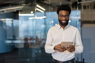 Başarılı ve gülümseyen bir adam pencerenin yanında tablet bilgisayarla duruyor, iş adamı sosyal ağlara bakıyor, eğitim materyalleri var. Ofis içi ofis çalışanı..