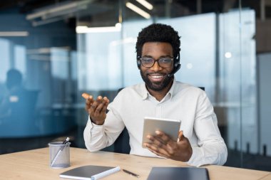 Kulaklıklı bir destek çalışanı kameraya bakar, elinde bir tablet bilgisayar tutar, tüketicilere uzaktan tavsiye verir ve yardım eder. Web kamerası görünümü.
