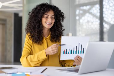 Çevrimiçi iş sunumu toplantısı. Mutlulukla gülümseyen bir kadın, finansal yatırım raporlarını paylaşıyor, grafiklerle çiziyor. İş yerindeki iş kadını video araması için defteri kullanıyor.