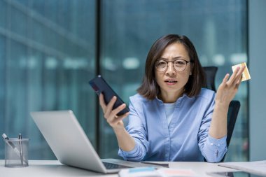 Kameraya bakan tatminsiz ve işkence görmüş Asyalı kadın. Ofis çalışanı elinde telefon ve banka kartı bulunan kameraya bakıyor. Hesap bloke edilmiş, hata ve dolandırıcılık..
