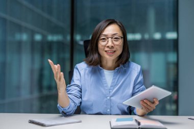 Tablet bilgisayarlı neşeli Asyalı kadın ofis masasında oturuyor. Gülümseyen ve kameraya bakan bir kadın, webcam görüntüsü, online sunum işi toplantısı.