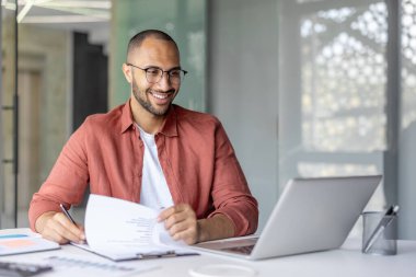 Ofis içi evrak işlerinde başarılı bir finansör. Sözleşmeler, faturalar ve raporlarla çalışan gülen adam. Vergi formlarını doldurmak için laptop kullanılıyor.