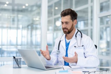 Klinikteki doktor video görüşmesi için dizüstü bilgisayar kullanıyor. Beyaz ceketli adam uzaktan konuşuyor. Ofisteki masada otururken hastalara danışıyor..