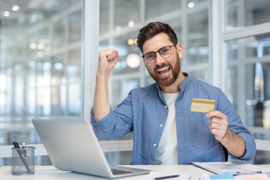 Banka kredi kartı olan bir adamın portresi, kameraya bakarak başarılı bir para transferini kutlayan bir çalışan, başarılı bir iş adamı, dizüstü bilgisayarla elini kaldırıyor..