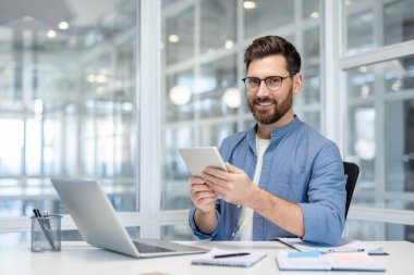 Başarılı genç bir iş adamının portresi, bir adam gülümsüyor ve elinde tablet bir bilgisayarla kameraya bakıyor, iş yerinde bir dizüstü bilgisayar kullanıyor, ofisinde çalışıyor..