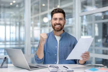 Başarılı yatırımcı başarının başarılı sonuçlarını kutluyor. Adam raporları elinde tutuyor ve zafer jesti olarak elini kaldırıyor. İş adamı kameraya bakar, ofis içinde dizüstü bilgisayarla çalışır..