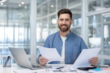 Kağıt işlerinde başarılı bir finansörün portresi, kontratları ve raporları olan, gülümseyen ve kameraya bakan bir adam, iş adamı neşeli bir yatırımcıdır..