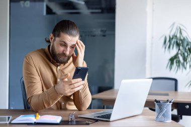 Profesyonel ofis ortamında çalışan bir adam akıllı telefonuna bakarken hayal kırıklığına uğramış görünür. Duygu, teknolojiyle ilgili stres ya da kafa karışıklığı taşır. Görüntü bir dizüstü bilgisayar içeriyor