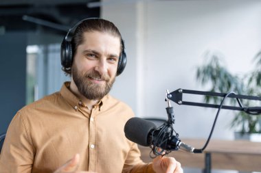 Masada oturan, kulaklık takan ve stüdyoda profesyonel bir mikrofona konuşan bir erkek podcast sunucusu. Gülümsüyor ve kendinden emin.