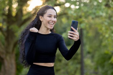 Joyful Hispanic woman outdoors, winning a challenge on her phone, pumps fist in excitement, smiling broadly. clipart