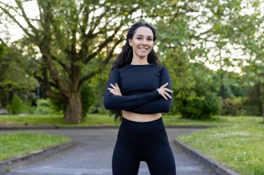 Neşeli, atletik kıyafetli bir İspanyol kadın, sağlıklı ve zinde bir yaşam tarzını sergilemek için lüks bir parkta çapraz kollarla poz veriyor..