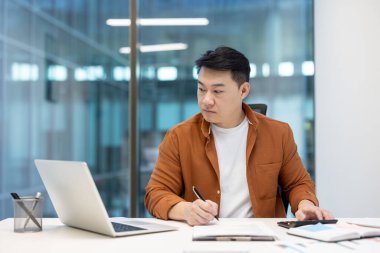 Ofis içinde evrak işleriyle uğraşan ciddi bir Asyalı finansör, hesap makineli iş adamı, ofis içi mali raporları ve vergi formlarını dizüstü bilgisayarla dolduruyor..