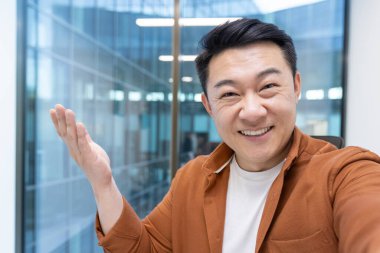 Mutlu ve başarılı Asyalı adam telefonda video uygulaması kullanarak konuşuyor. Ofiste çalışan bir adam, kameraya bakıyor..