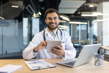 Ofisteki başarılı deneyimli doktor elinde dizüstü bilgisayarla masada oturuyor. Elinde tablet bilgisayarla beyaz önlüklü bir adam kameraya bakar ve gülümser..