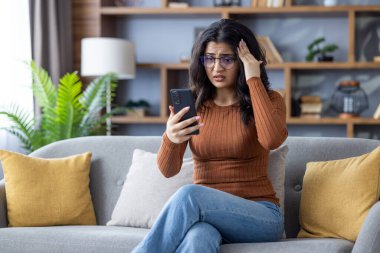 Şok olmuş genç Hintli kadın evinde koltukta oturuyor, telefon ekranına bakıyor, el ele tutuşuyor..