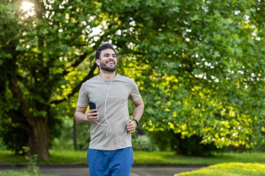 Adam telefon ve kulaklıkla aktif fiziksel egzersiz ve park yerinde spor yapıyor. Yeşillikte koşmanın keyfine varır, sağlığını ve sağlıklı yaşam tarzını destekler..