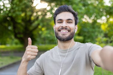 Kulaklık takan, gülümseyen ve elinde tuttuğu kameraya bakan genç Hintli bir adamın portresi..