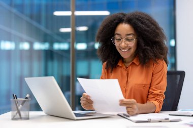 Başarılı kadın finansör elinde bir rapor tutuyor, okuyor ve gülümsüyor. İş kadını ofisin içindeki işyerindeki çalışmalarının sonuçlarından memnun..
