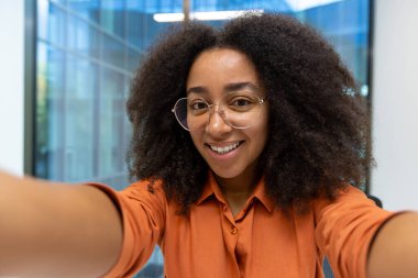 Webcam görüntüsü, ofisin içinde fotoğraf çeken genç bir kadın. Kadın çalışan kameraya bakıp gülümsüyor, akıllı telefondaki video arama uygulamasını kullanıyor..