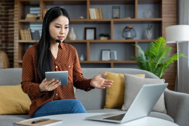 Asyalı kadın video araması için laptop ve tablet kullanıyor. Kulaklık takıyor, ev ofisinden uzaktan çalışıyor. Uzaktan çalışma, sanal iletişim ve modern teknolojiyi içerir..