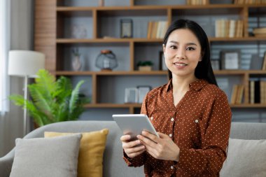 Modern evdeki kanepede tablet tutan Asyalı kadın. Gülümseyen, rahat, rahat, şık oturma odası dekorunun keyfini çıkaran. Ev dekorasyonu barışçıl havayı arttırır. Teknoloji ve yaşam tarzı kavramı.