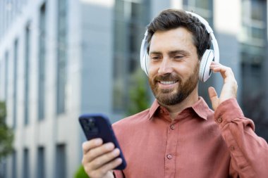 Bir adam açık havada kulaklıkları ve akıllı telefonuyla en sevdiği müziği dinlerken mutlu bir şekilde gülümsüyor..