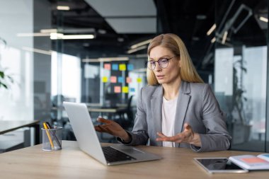 Ciddi odaklı iş kadını bilgisayardaki video görüşmesini kullanarak iş arkadaşlarıyla uzaktan konuşuyor. Ofis içi iş elbiseli kadın, online konferans toplantısında..