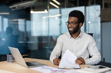 Ofis içinde evrak işlerini yapan başarılı genç bir finansör. Adam mutlu bir şekilde gülümsüyor, dizüstü bilgisayarla, belgelerle, grafik ve çizelgelerle çalışıyor. İş adamı gömleği hazırlıyor..