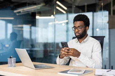 Telefondaki uygulamayı kullanan ciddi odaklı bir adam. İşadamı, ofisin ortasında masada oturup, akıllı telefondan mesaj yazmayı ve internet sayfalarına göz atmayı düşünür..