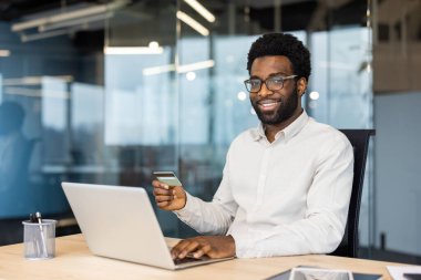 Banka kredi kartıyla kameraya bakan neşeli ve başarılı bir adam. İşyerindeki işadamlarının portresi, çalışanların internet üzerinden para transferi ve kitap hizmetleri