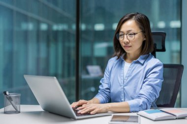 İş yerinde bilgisayarla çalışan kendine güvenen Asyalı bir kadın. Klavyede yazan kadın, elektronik rapor hazırlıyor. Masada oturan düşünceli ve konsantre genç iş kadını..