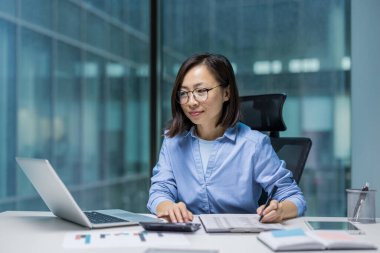 Ofis içinde evrak işi yapan ciddi odaklı Asyalı kadın. İş kadını vergi formlarını ve mali raporları dolduruyor. İş yerinde bilgisayarını düşünceli bir şekilde kullanan kadın..