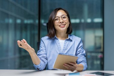 Webcam görüntüsü, hareket halindeki bir defteri tutan, gülümseyen ve kameraya bakan bir kadın. Çevrimiçi iş konferansındaki Asyalı kadın, verileri kaydediyor ve işyerinde ofis içi eğitim görüyor..