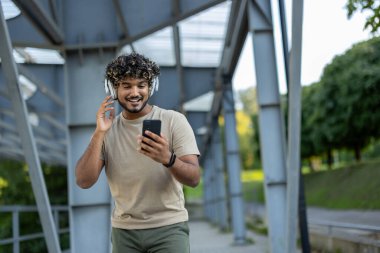 Spor kıyafetli genç adam parkta yürüyor. Telefonunda müzik dinlemek için kulaklık ve uygulama kullanıyor. Ses yayını ve sesli yayınlar..