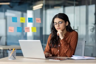 Ofis içinde dizüstü bilgisayarı olan ciddi düşünen bir kadın. İş kadını oturuyor ve ekrandan veri okuyor. Çalışan özenle güncel sorunları çözüyor.