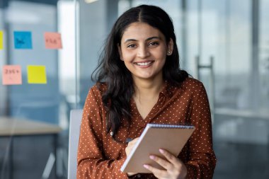 Webcam görüntüsü, elinde not defteri tutan bir kadın. İş toplantısı, çevrimiçi konferans, çalışanlar ofiste otururken mutlu bir şekilde veri yazıyorlar..